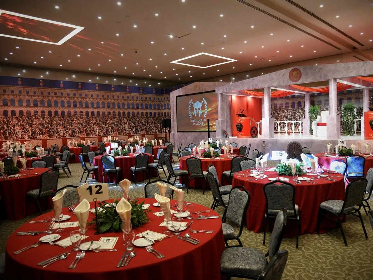 Um salão de gala preparado para um jantar de showcase, com mesas vermelhas e um ecrã no palco